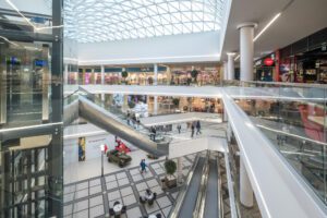 Interior view of a retail space with escalators, stores, and a large skylight overhead.