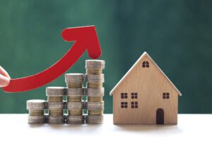 A red arrow ascends above coin stacks next to a wooden residential property model, symbolizing the rise in housing costs or investment value.
