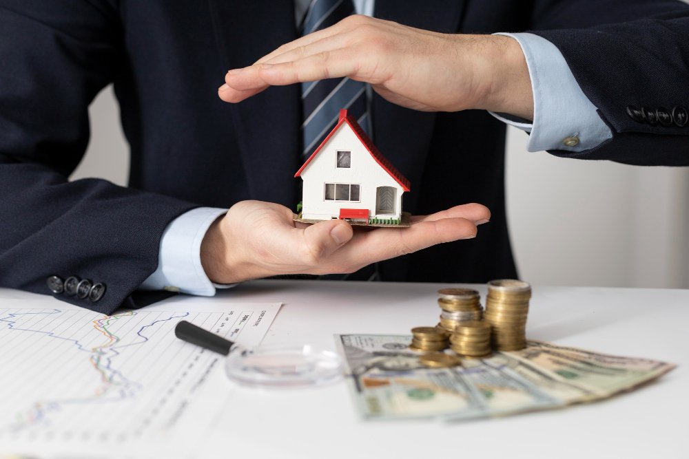 A professional in a suit protectively covers a model house, illustrating safeguarding investments from real estate scams, with coins and cash displayed.