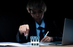 A person in a suit holding a key above a small model house, with a laptop and documents in the background, highlighting the concept of mortgage fraud.